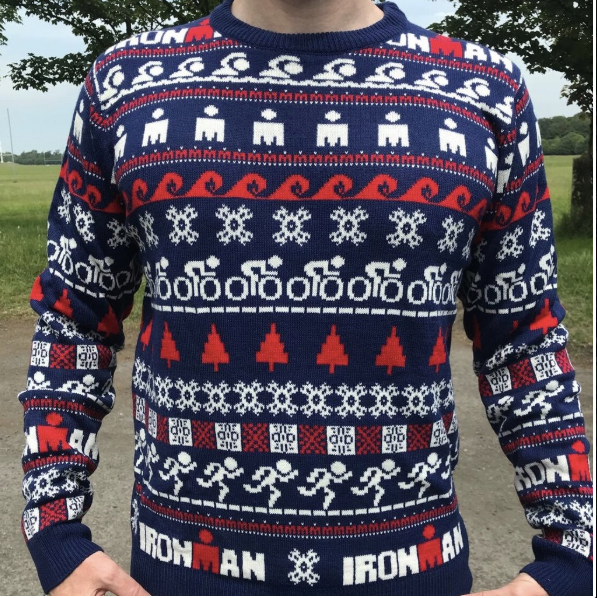 Person wearing a navy custom IRONMAN knitted Christmas sweater by Roody, with a red-and-white Fair Isle pattern of swimmers, cyclists, and runners and "IRONMAN" knitted into the design, photographed outdoors with trees in the background.