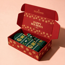Load image into Gallery viewer, An open red gift box with gold holiday patterns and the words 'HAPPY HOLIDAYS from Brand' inside, containing a pair of green knit socks with gold and white festive patterns, set against a solid pink background.