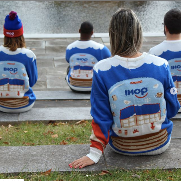 Group of people seen from behind, sitting on outdoor stone steps and wearing matching royal-blue custom IHOP knitted Christmas sweaters by Roody, with the IHOP pancake-stack logo on the back.