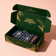 Load image into Gallery viewer, An open dark green gift box with gold holiday illustrations and the words 'Happy Holidays' inside, containing a folded navy knit sock with white and orange holiday patterns. The box is photographed on a soft peach-pink background.
